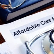 A close-up of paperwork with Affordable Care Act printed at the top of it. A stethoscope resting on top of the paperwork & a book w/a pair of glasses resting on top.